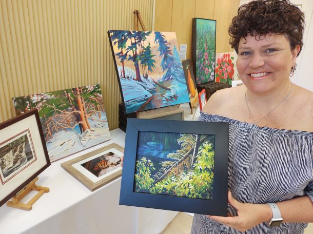 Image shows a smiling woman indoors holding a small framed painting that depicts an outdoor forest and rive scene. There is more artwork in the room behind her.