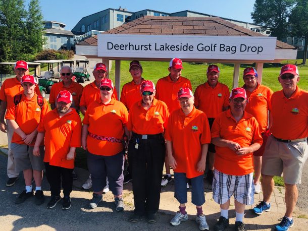 Image shows a group of 14 smiling people in matching orange T-shirts and red hats standing outside. A sign behind them reads: Deerhurst Lakeside Golf Bag Drop.