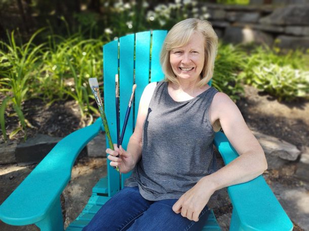 Image shows a smiling woman holding three fine art paintbrushes, while sitting in a blue Muskoka chair outside in a garden.