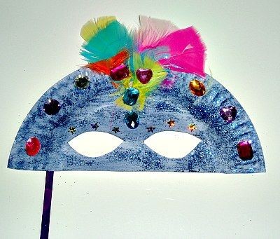 Image shows a colourful mask made from half a paper plate. It is meant to cover the top of the face. It has two eye holes cut out of it. It is painted blue and has plastic jewels and feathers glued to it. 