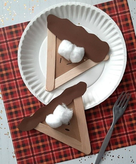 Image shows a craft that looks like a wedge of pecan pie made from a paper plate, brown construction paper, popsicle sticks and cotton balls atop a red checkered placemat.