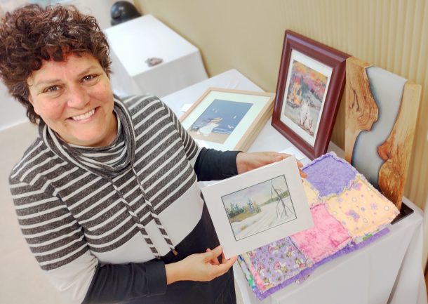 A smiling woman looks up at the camera while holding a small, unframed winter landscape painting. Other art items are on a table beside her, including a colourful fabric lap quilt, a wood and resin sculpture and two other framed pieces of art.