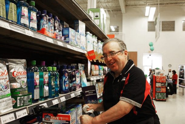 A man in a grocery store aisle smiles at the camera while placing products on the shelf.