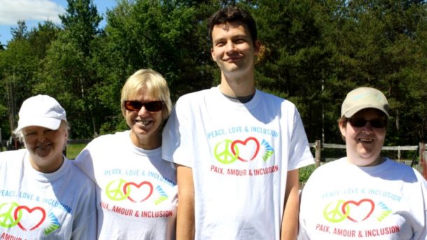 Four people stand together in white T-shirts that read: Peace Love and Inclusion