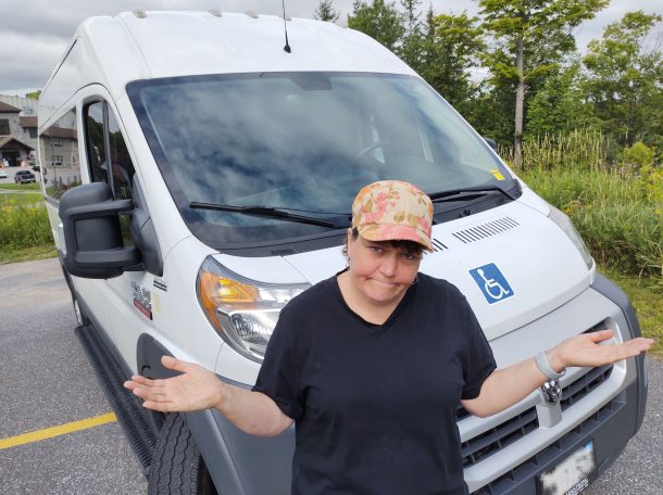 Woman in front of large white van stands with arms out and hands palms up, as if to say, 'Would you want to ride in this?'