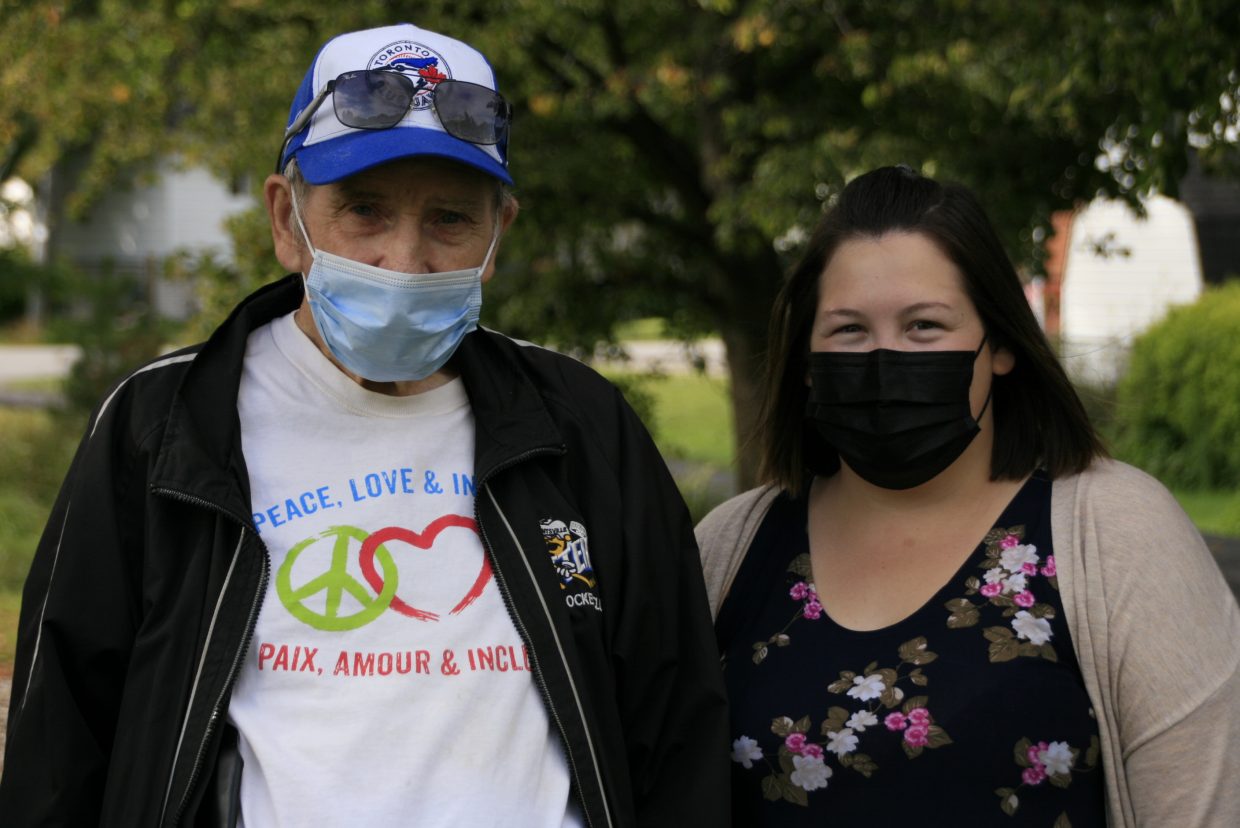 A man and a woman wearing face masks pose for the camera. They are standing near a tree in a park. The man's shirt reads: Peace, Love, and Inclusion. Paix, Amour, and Inclusion.