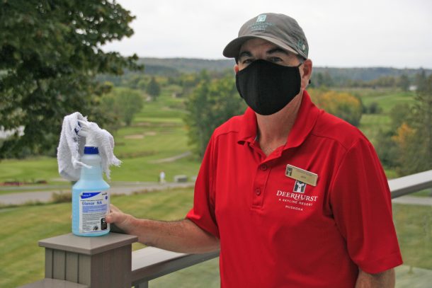 A man in a red golf shirt and black face mask stands on an outdoor patio. There is a golf course behind him. He wears a name tag and holds a bottle of window cleaner. Text reads: Jeff. Deehurst Muskoka.