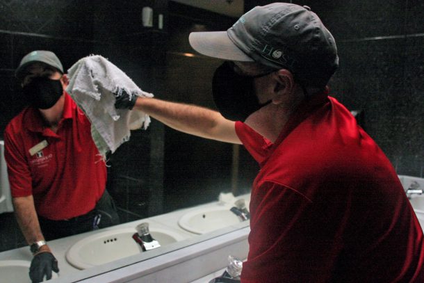 A man in a red shirt and black face masks wipes clean a bathroom mirror using a white cloth.