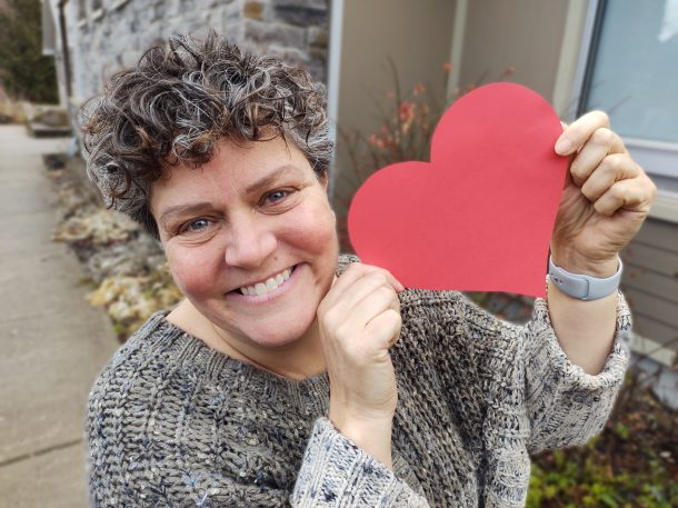 A photo of a smiling woman holding a red piece of paper in the shape of a heart.