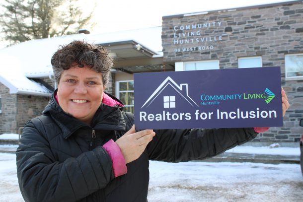 A photo of a smiling woman standing outside while holding a small, dark blue sign. Sign reads: Community Living Huntsville Realtors for Inclusion.