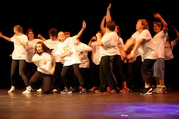 A photo of many people dancing as a group on a theatre stage. Everyone wears a white T-shirt and black pants. The shirts have colourful hearts on them and read: Peace, Love and Inclusion.