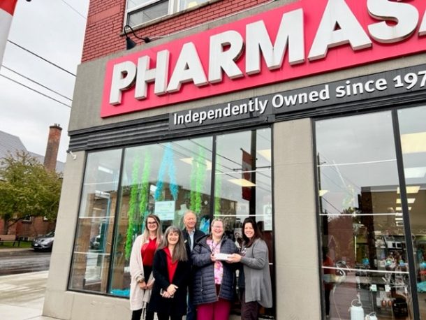 5 people stand outside in front of a Pharmasave Huntsville window that has blue and green feather boas and other decorations on display. 1 person hands a box of baked goods to another. Everyone is smiling.