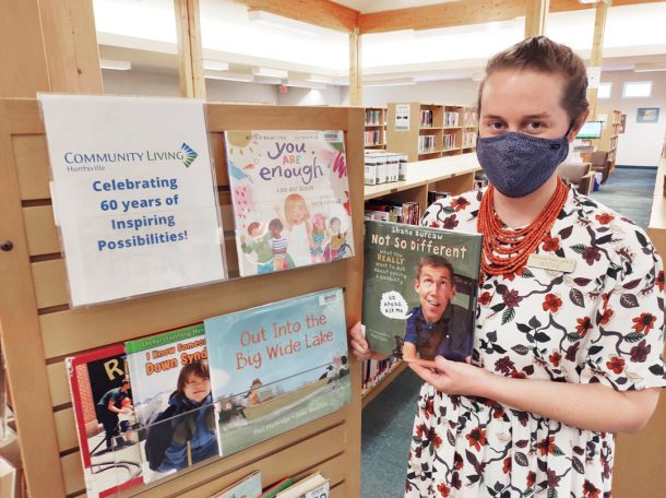 A woman in a cloth face mask and floral print dress holding a book near a full bookshelf. Sign reads: Community Living Huntsville, celebrating 60 years of inspiring possibilities.