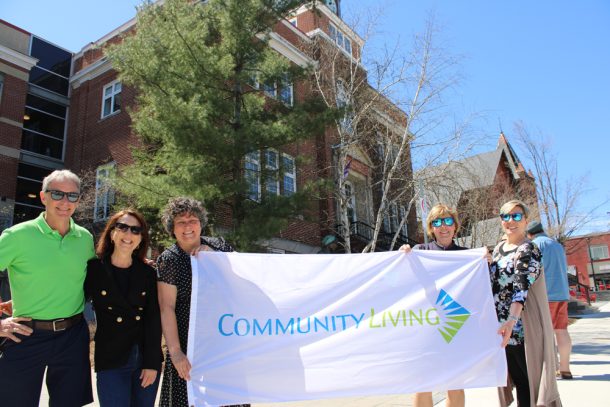 5 people hold a large Community Living flag with Huntsville town hall behind them.