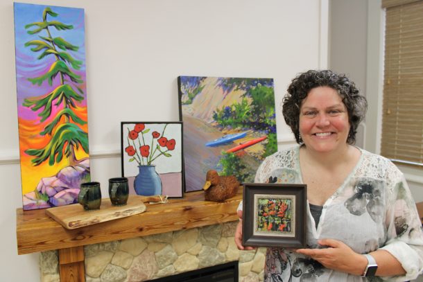 A smiling woman stands indoors and holds a small framed landscape painting while standing in front of a faux fireplace. On the fireplace mantle sits three paintings, 2 ceramic mugs, a handcrafted wood cutting board, and a whittled wooden duck.