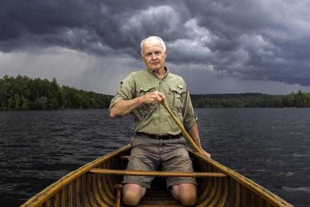 A man, Roy MacGregor, kneels in a canoe and paddles on an Algonquin Park lake. The sky is dark, cloudy, and ominous.