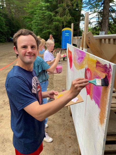 A young man smiles for the camera as he paints on a large outdoor canvas.