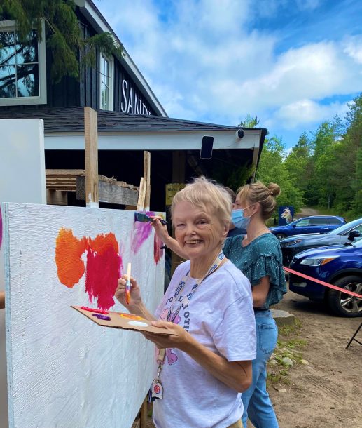 A woman smiles as she paints on a large outdoor canvas.