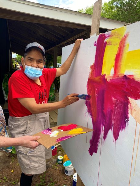 A man in a red T-shirt pauses for a photo while he paints a large outdoor canvas.