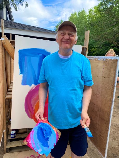 A man in a blue T-shirt smiles for the camera. Behind him, there is a canvas with blue and purple paint on it.