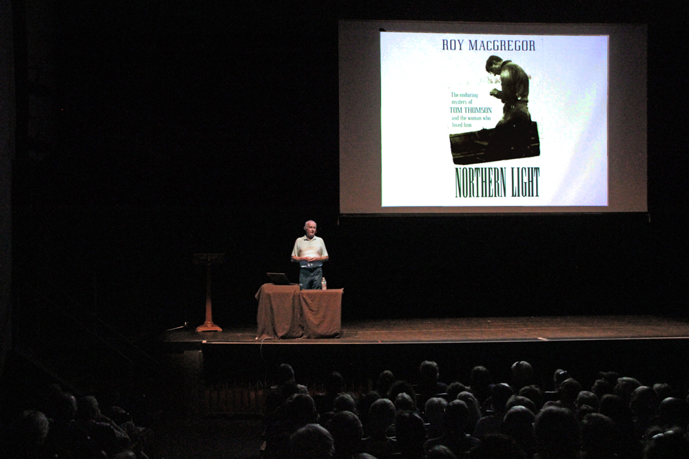 Roy MacGregor on stage at the Algonquin Theatre. A projection screen with the cover of his book, Northern Light, is behind him. Audience members can be seen in the seats below him.