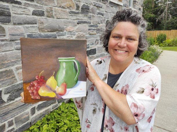 A smiling woman holds a still life painting of a green jug, grapes, and pears, while standing outside in front of a stone wall.