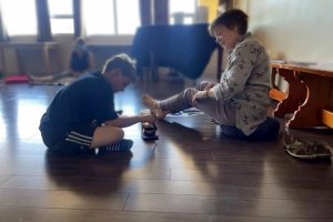 A boy sits cross-legged on the floor while sounding a singing bowl with help from an adult facilitator.