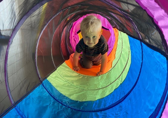 A smiling child crawls through a colourful fabric tunnel.