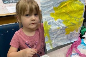 A young girl holds up her colourful blue and yellow painting.