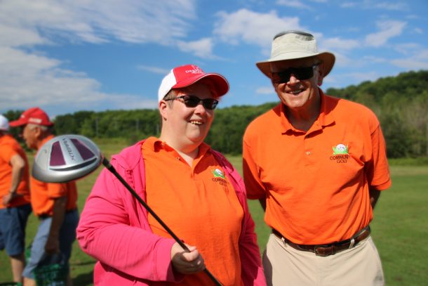 A woman in an orange shirt, pink sweater and pink and white ball cap holds a golf club while standing beside a man in an orange shirt and white broad-rimmed hat. They are on a golf course.