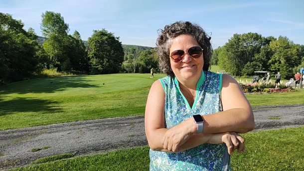 A smiling woman in sunglasses and a sleeveless green and blue golf shirt leans on a golf club while standing on a golf course on a sunny day.