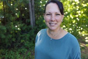 A smiling woman in a blue top poses for a photo outdoors. There are trees with green leaves behind her.