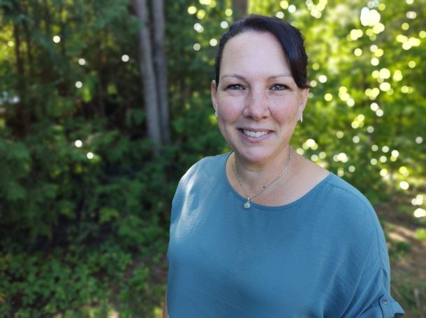 A smiling woman in a blue top poses for a photo outdoors. There are trees with green leaves behind her.