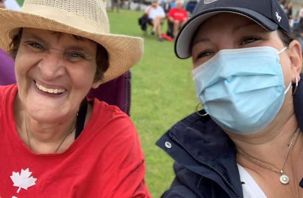 A smiling woman in a wide-brimmed hat and red T-shirt takes a selfie at at a community festival with a woman in a ball cap, face mask and blue jacket.
