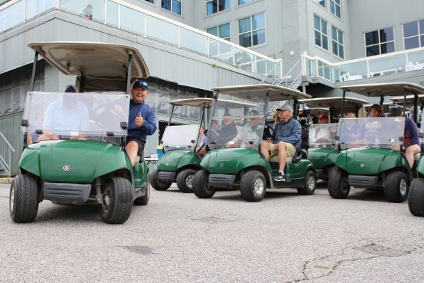Green golf carts filled with people head out from a golf course clubhouse. A man in the first cart leans out and gives a thumbs up.