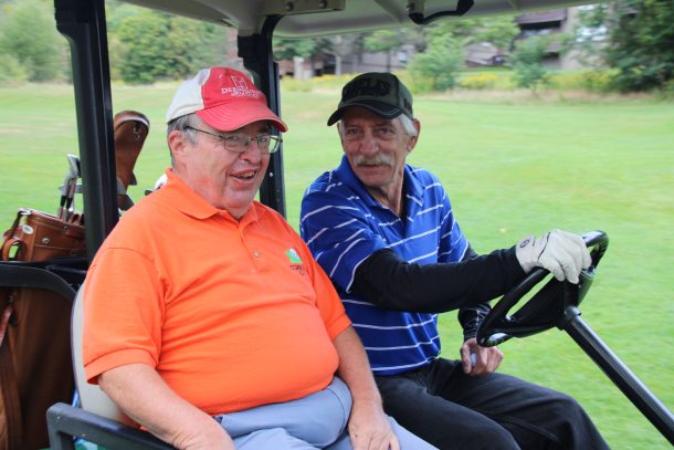 2 men smile for the camera while riding in a golf cart.