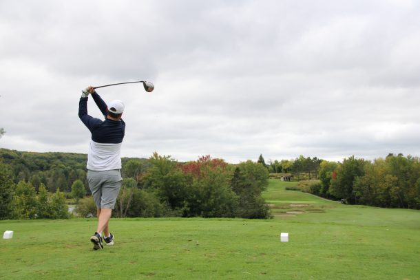 A man swings through a tee off while on a green golf course surrounded by trees.