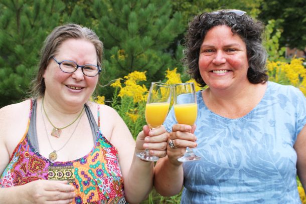 2 smiling women clink glasses of orange juice while standing outside surrounded by green trees and golden rod plants.