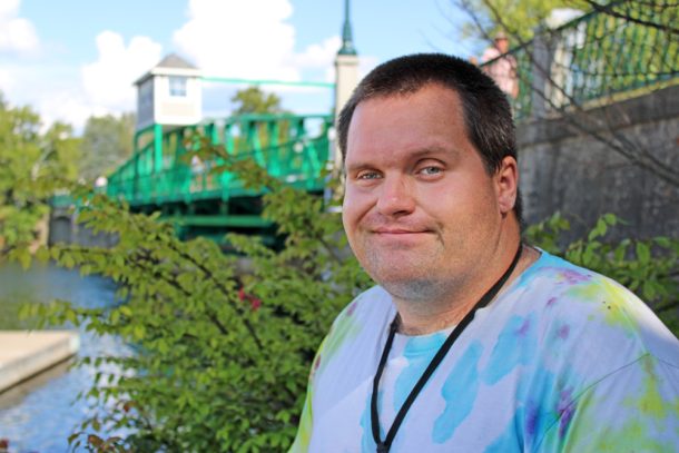 A smiling man with short hair and a white tie dyed T shirt stands outside with a green shrub and green metal bridge in the background behind him.