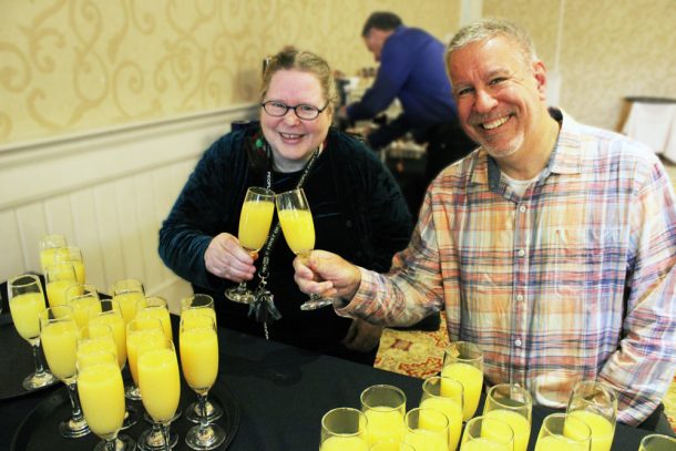 A woman and man smiling while clinking glasses of orange juice.