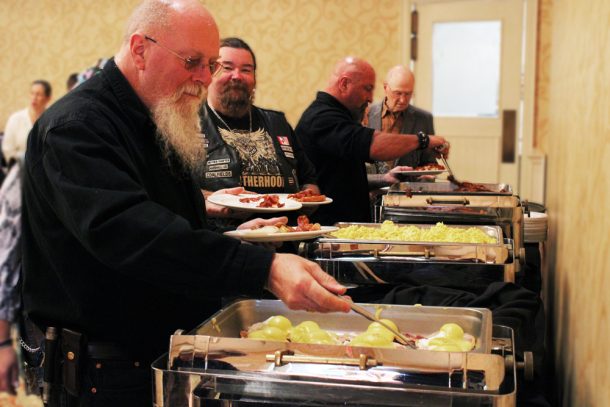 People serving themselves from a breakfast buffet. Man closest to the camera smiles as he helps himself to an eggs benedict.
