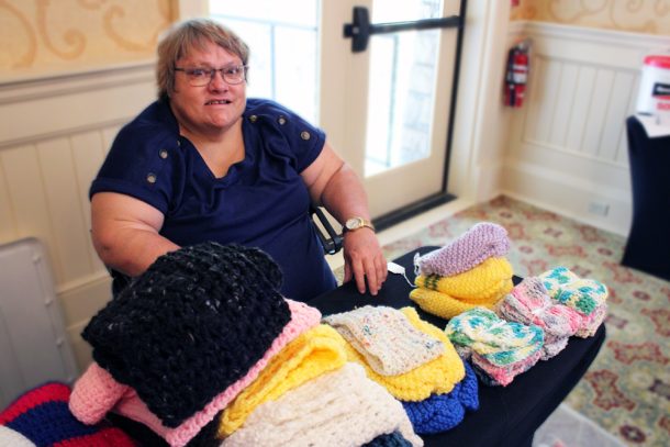 A woman smiles while seated at a table filled with knitted hats and cloths.