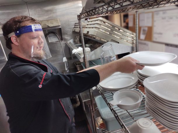 A man in a black chef jacket and face shield stacks clean plates in a restaurant kitchen.