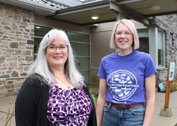 2 smiling women stand outside a grey-stone building. 1 has long grey hair and a purple top, and the other has short blonde hair and a blue T-shirt.