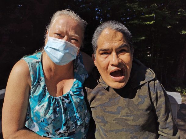 A woman with blonde hair, blue shirt, and blue face mask sits outside with a smiling man with grey hair and brown sweater. There are trees in the background.