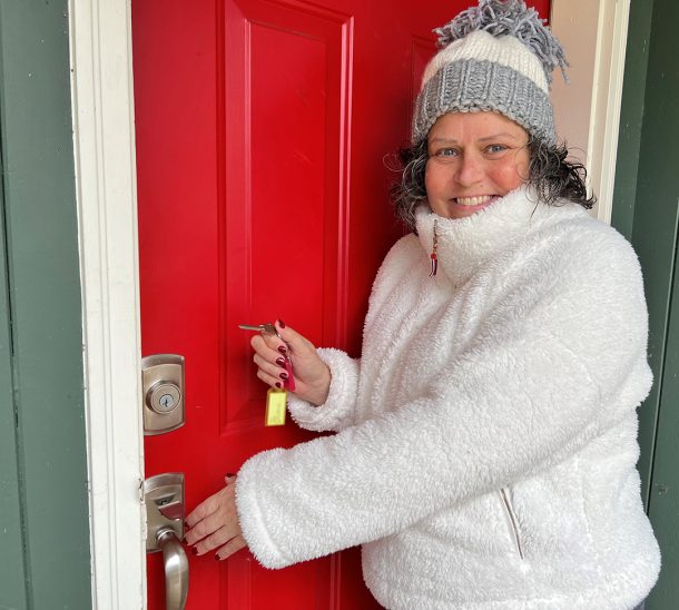 A smiling woman in a toque and winter coat holds a house key while reaching for the nob of a house's front door.