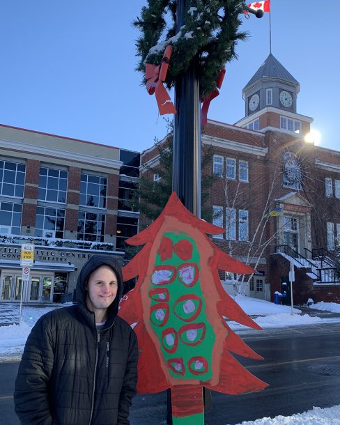 Man in winter coat and hat on sidewalk near lamp post with decorative wooden Christmas tree attached. Tree has painted green centre with red circles and branches.