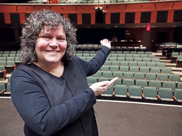 A smiling woman, Jennifer, stands on a theatre stage and gestures with her hands to the empty theatre seats behind her.