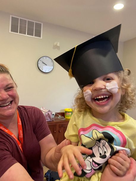 A laughing young girl in a yellow T shirt and paper graduation cap with a laughing adult.