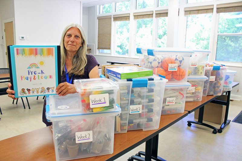 A woman, Yvonne, holds a Pre K Play and Learn binder while standing beside clear tote boxes filled with program supplies.